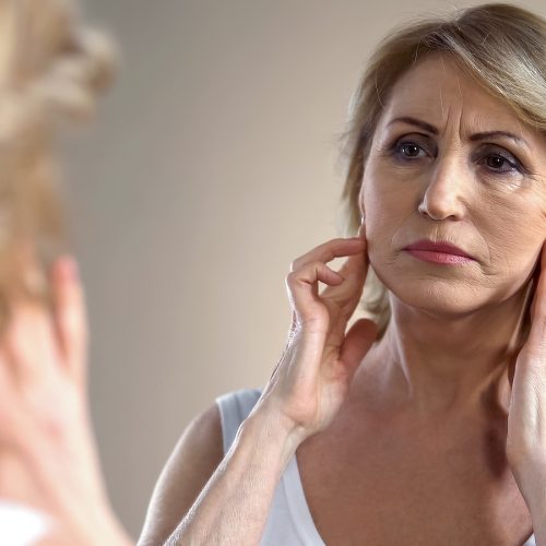 Unhappy aged woman looking in mirror at home, touching face, aging process